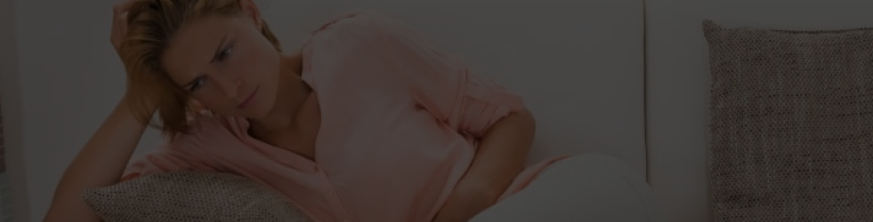 Woman lying on couch with one hand on her head and one on her belly — showing signs of hormonal imbalance, stress, and irritability for desktop view
