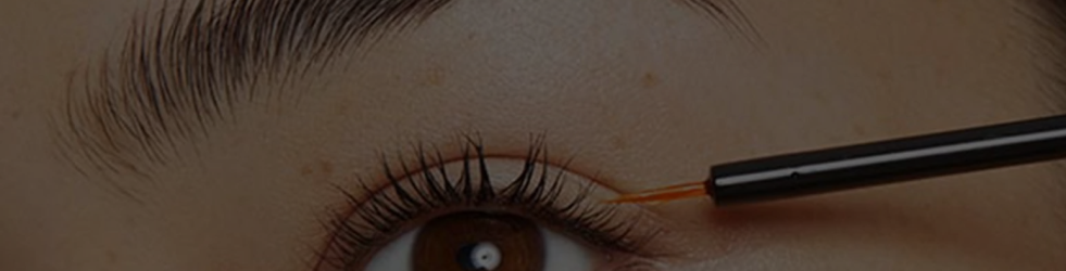 Woman applying lash oil to her eyelashes using a black applicator wand with light brown bristles — showing close-up lash care and growth for desktop display
